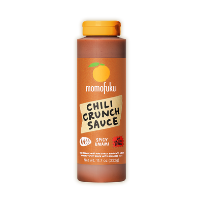 A bottle of Momofuku Chili Crunch Sauce in Spicy Umami flavor, featuring a brown label with an orange cap, shown on a plain white background.