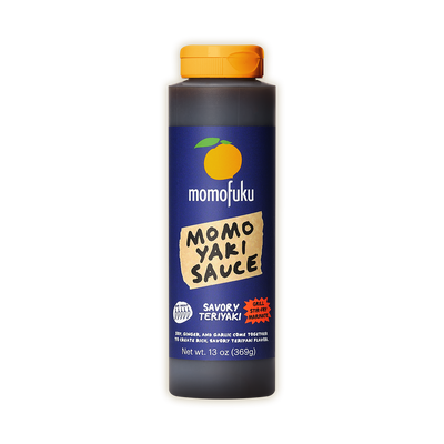 A bottle of Momofuku Momo Yaki Sauce on a plain white background. The bottle is dark gray with a bright orange cap and a navy blue label featuring an orange graphic and a beige paper-style label.