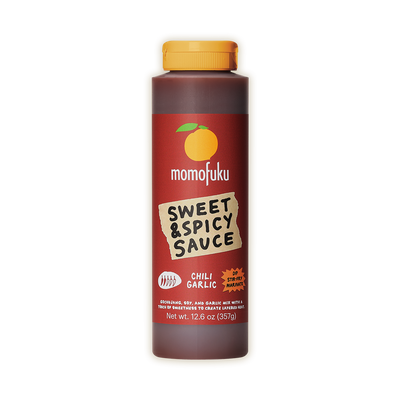 A bottle of Momofuku Sweet & Spicy Sauce in Chili Garlic flavor, featuring a dark reddish-brown label with a yellow-orange cap, set against a plain white background.