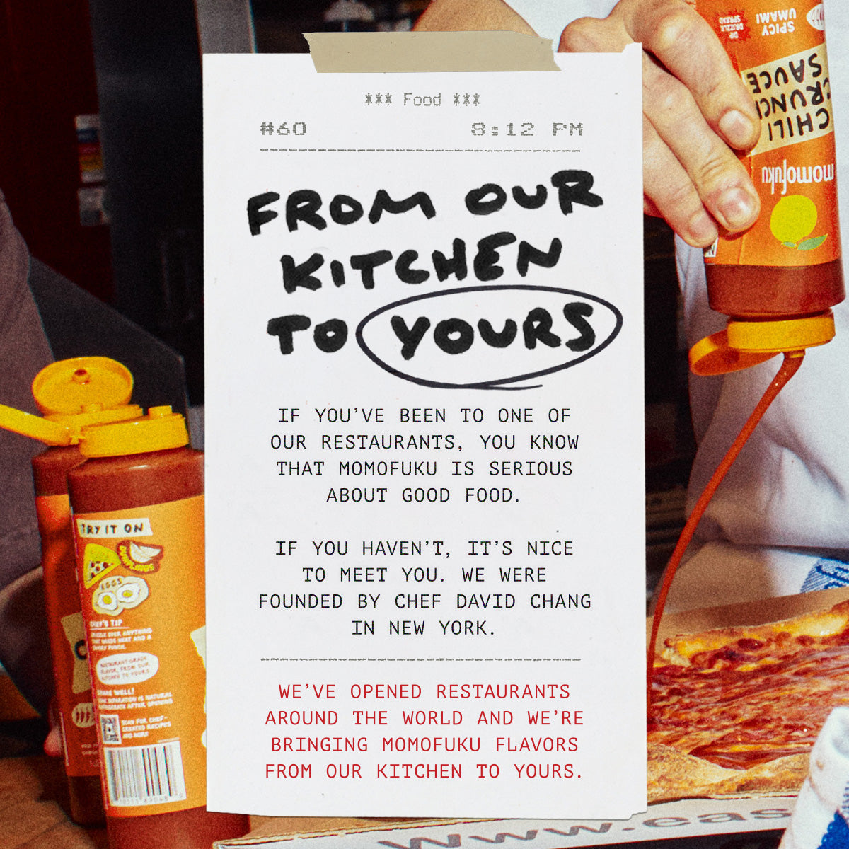 A printed paper menu or flyer held in a restaurant setting, with a hand squeezing a bottle of chili oil sauce onto it. The flyer reads 'FROM OUR KITCHEN TO YOURS' and includes text about Momofuku and restaurant origins.