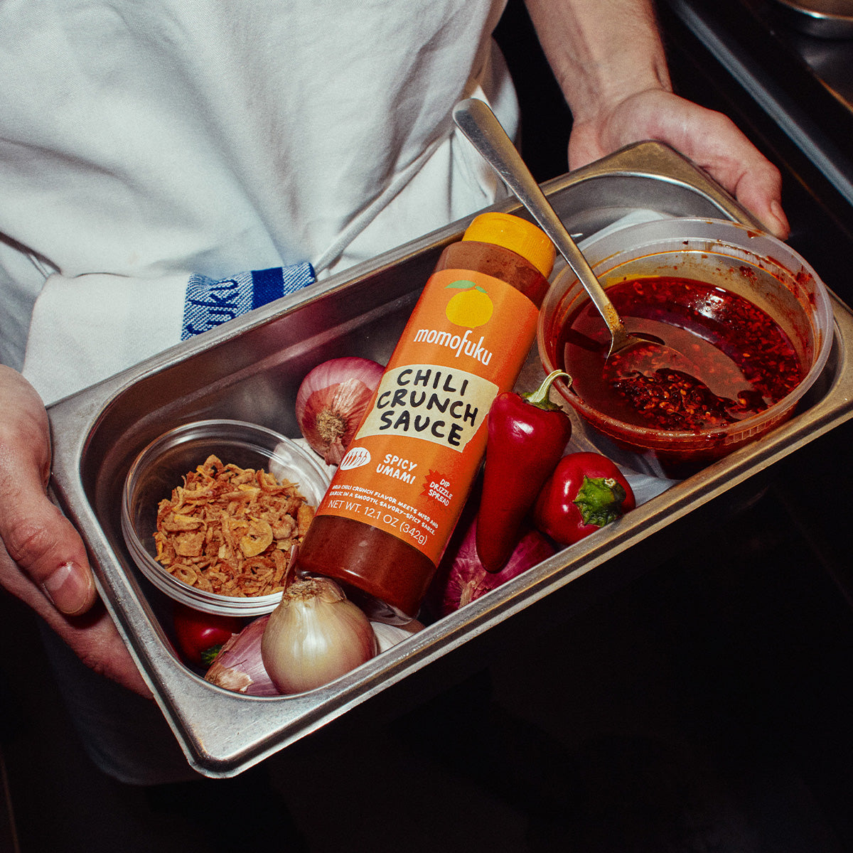 A stainless steel food prep tray held by a person wearing a white chef's coat, containing Momofuku Chili Crunch Sauce, red bell peppers, onions, and a bowl of red sauce with a spoon.