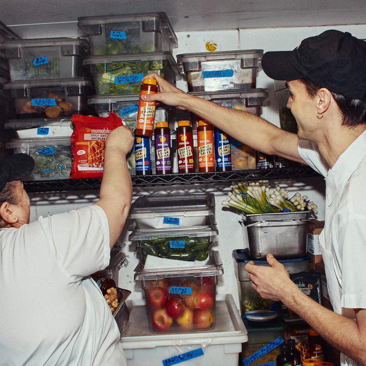 Two people in white shirts and black caps reach into a refrigerator shelf stocked with bottled beverages and plastic food storage containers filled with produce.