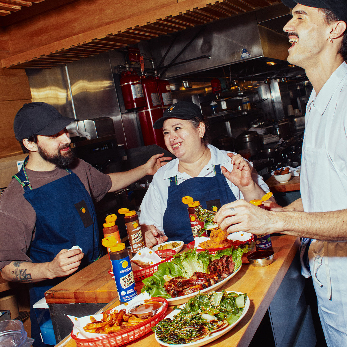 Three people in navy blue aprons smiling and laughing in a commercial kitchen, with plates of food and condiment bottles on a wooden counter in the foreground.