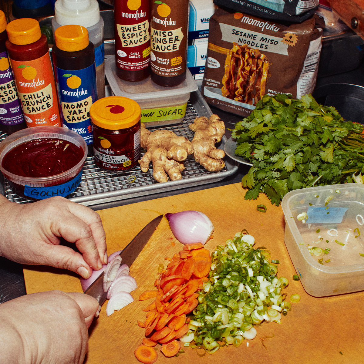 A person's hands slicing a red onion on a wooden cutting board, surrounded by chopped carrots, scallions, cilantro, and various Momofuku sauce bottles and a box of miso noodles in a kitchen setting.