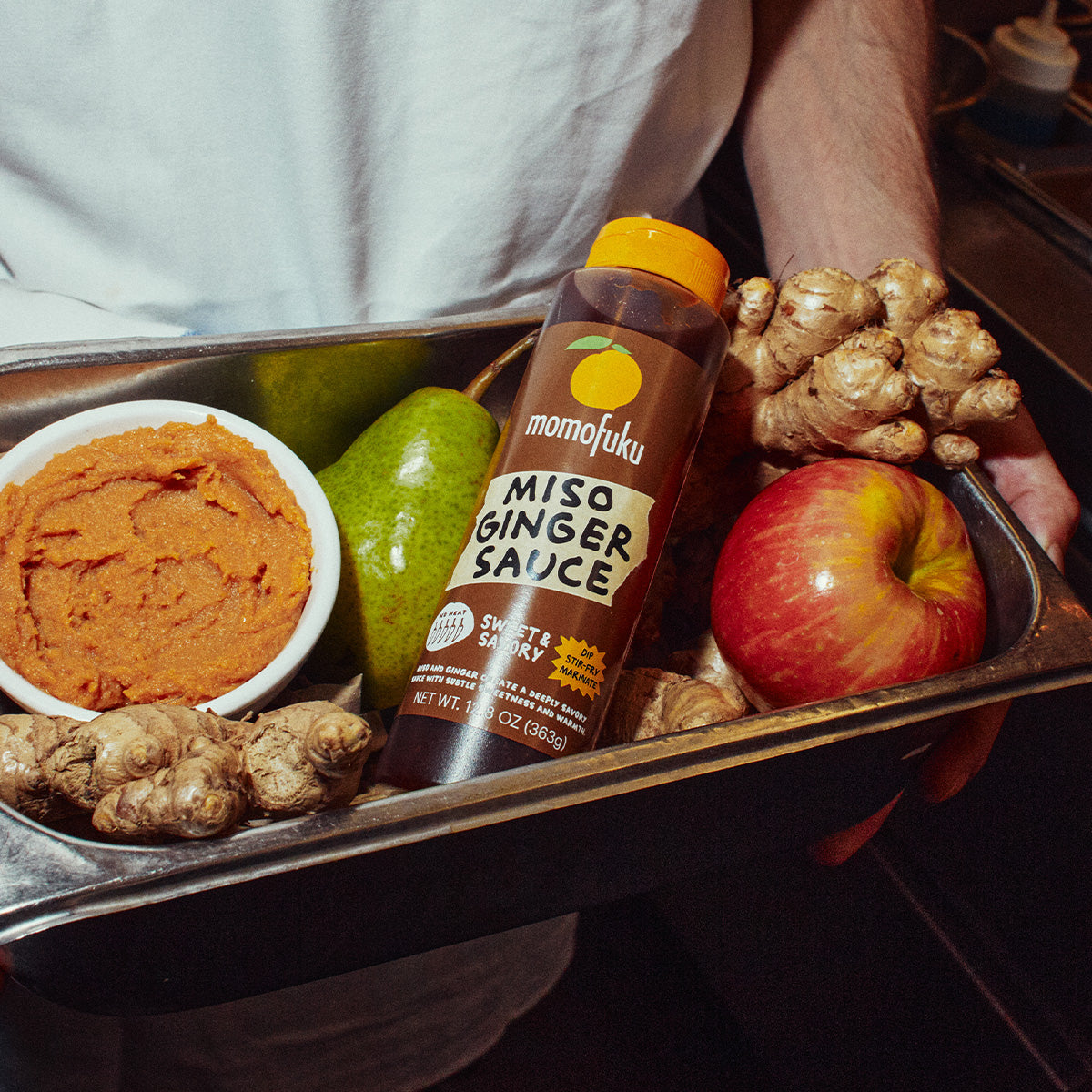 A person holds a metal tray containing a bottle of Momofuku Miso Ginger Sauce, along with ginger root, a bowl of orange-colored paste, a green pear, and a red apple in a dimly lit kitchen setting.