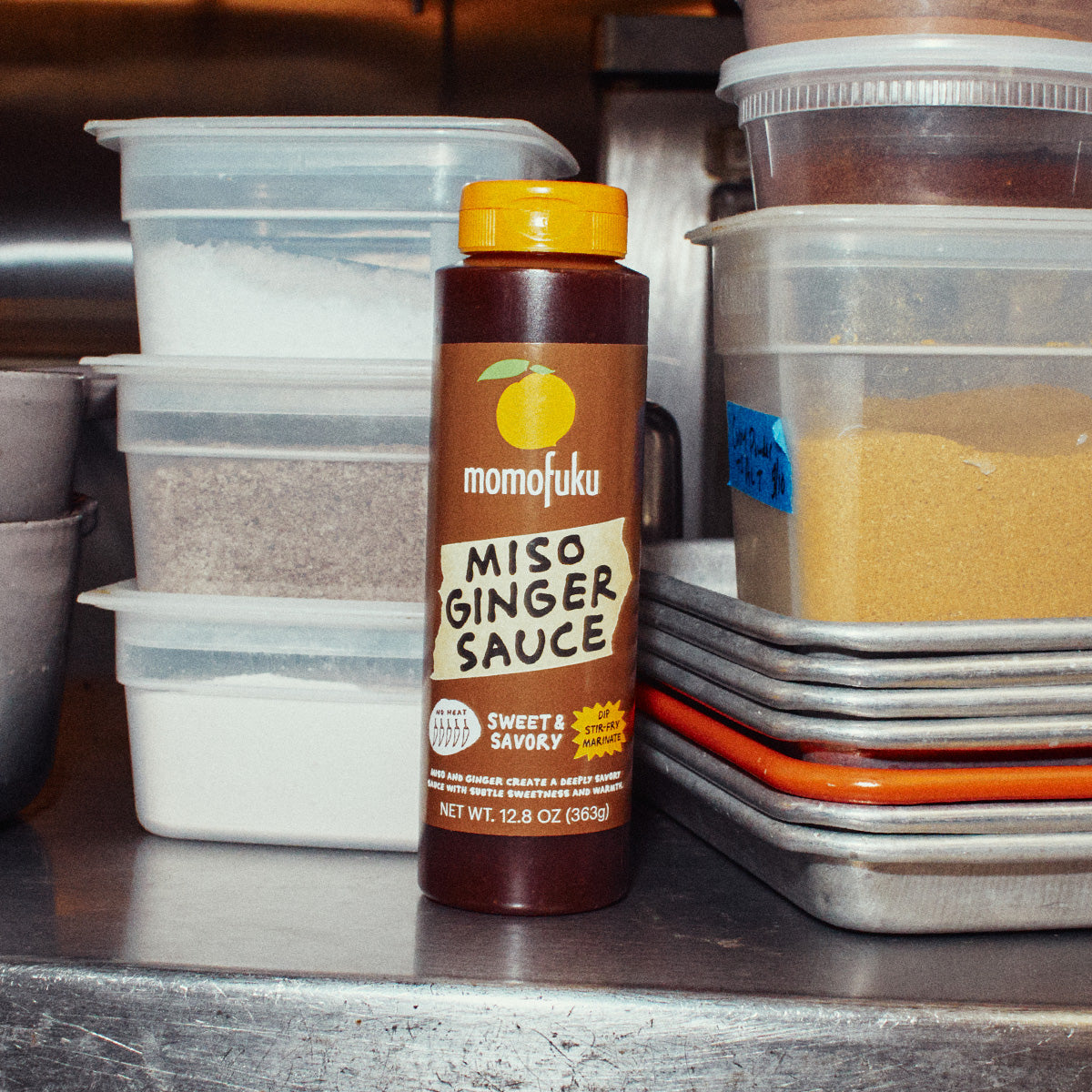 A bottle of Momofuku Miso Ginger Sauce stands on a stainless steel counter, surrounded by stacked plastic containers of ingredients in a kitchen setting.
