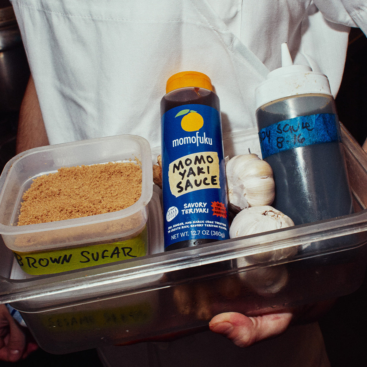 A person in a white chef's coat holds a plastic tray containing cooking ingredients and bottles, including a bottle of Momojuku Momoyaki Sauce, a squeeze bottle, garlic cloves, and a container of brown sugar.