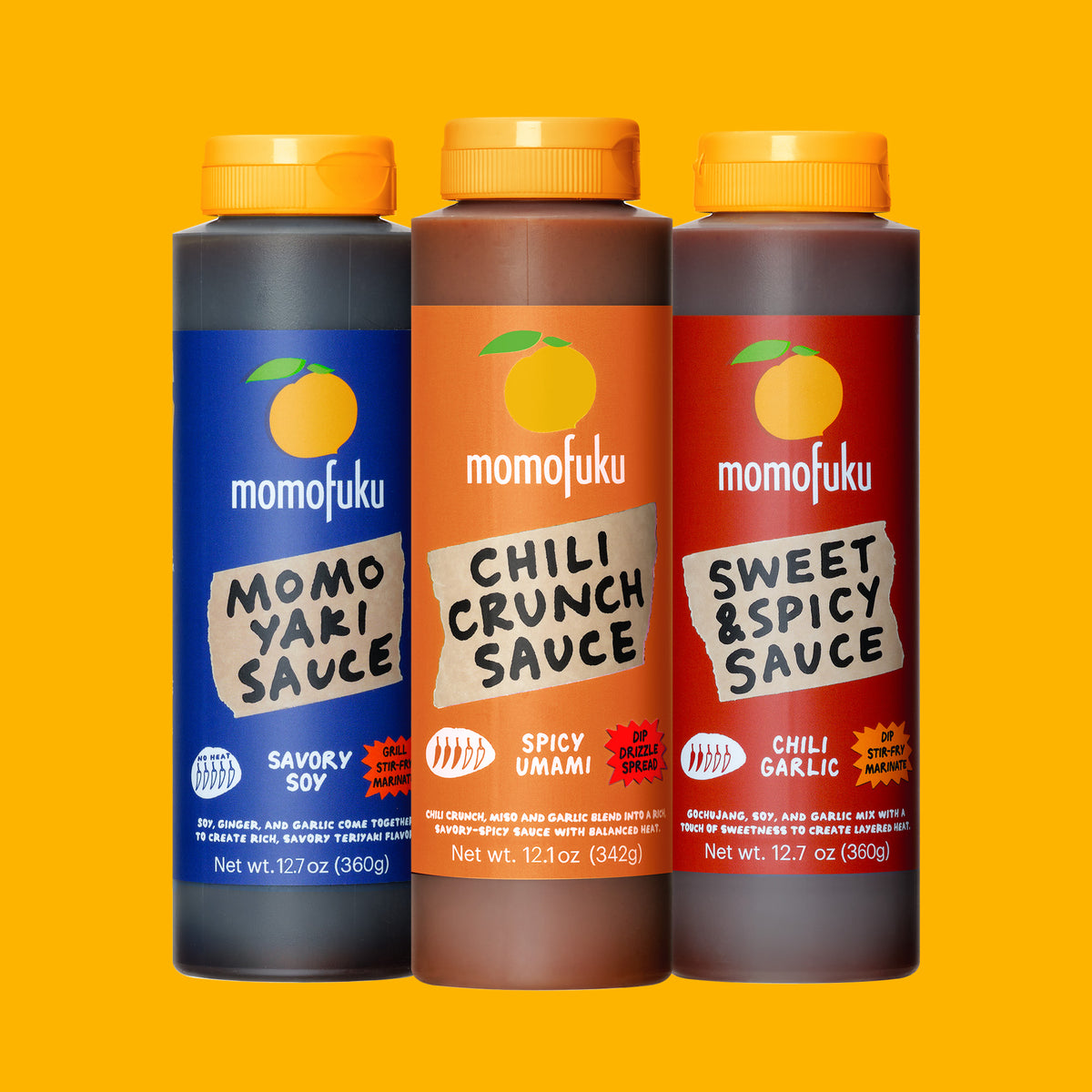 Three bottles of momofuku sauce lined up against a solid yellow background: Savory Soy, Spicy Umami, and Chili Garlic flavors. The bottles are dark gray, brown, and red respectively, with yellow caps and orange labels.