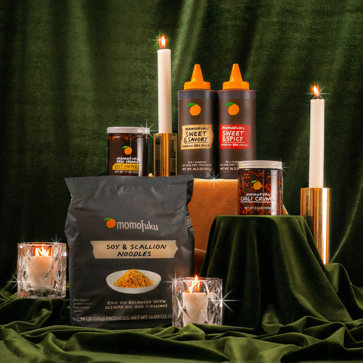 Festive display of Soy & Scallion Noodles, 1 jar Chili Crunch, 1 jar Chili Crunch Hot Honey,  1 Bottle of Sweet & Savory Korean BBQ Sauce, 1 Bottle Bottle of Sweet & Spicy Korean BBQ Sauce on velvet green background