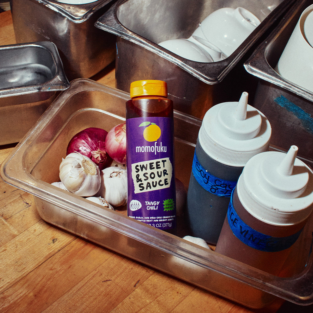 A bottle of Momajuku Sweet & Sour Sauce in Tangy Chili flavor, placed in a clear plastic container with garlic, red onion, and squeeze bottles on a wooden surface.