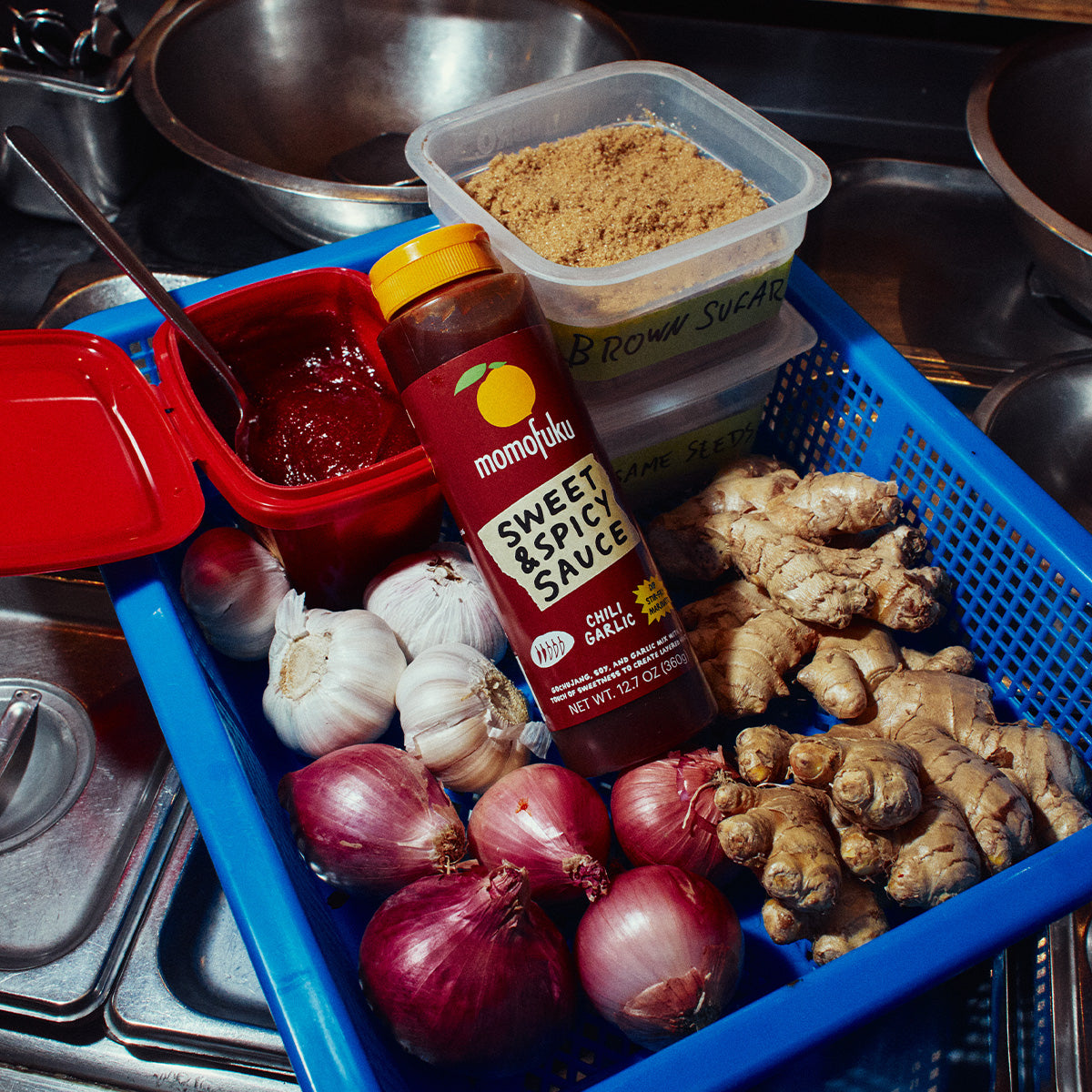 A blue plastic basket filled with cooking ingredients on a stainless steel kitchen sink, including a bottle of Mom's Ajita Sweet & Spicy Sauce, a red chili sauce container, brown sugar, red onions, garlic, ginger, and shallots.