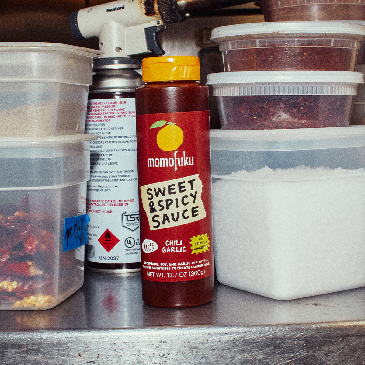 A bottle of Momofuku Sweet & Spicy Sauce (Chili Garlic) stands on a stainless steel counter surrounded by kitchen supplies, including plastic containers of ingredients and a propane torch.